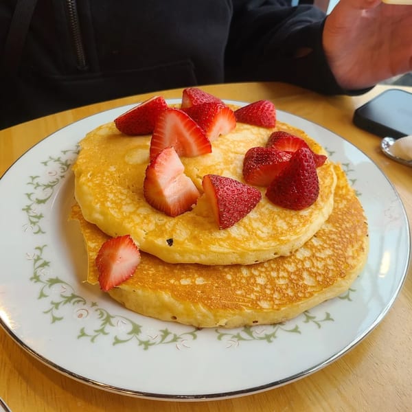 Stack of golden pancakes topped with fresh, sliced strawberries on a white plate with green floral trim.