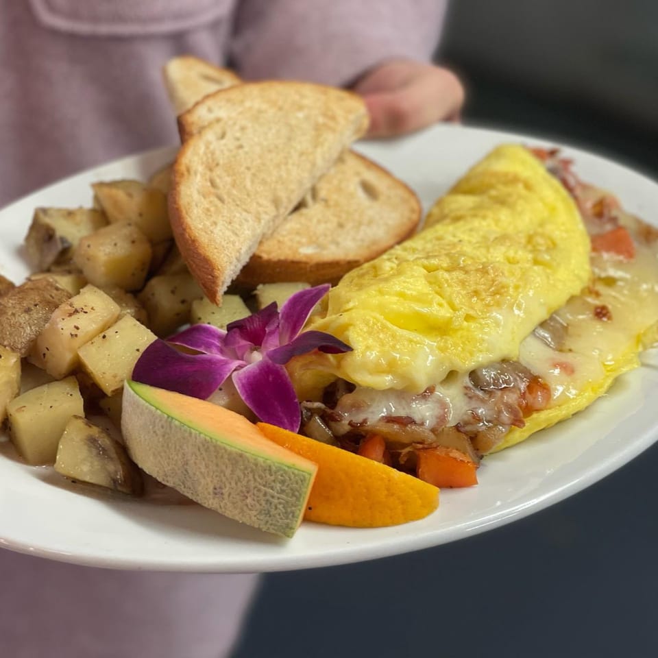 A breakfast plate with a cheesy omelet, home fries, toast, cantaloupe, orange slice, and a purple orchid.